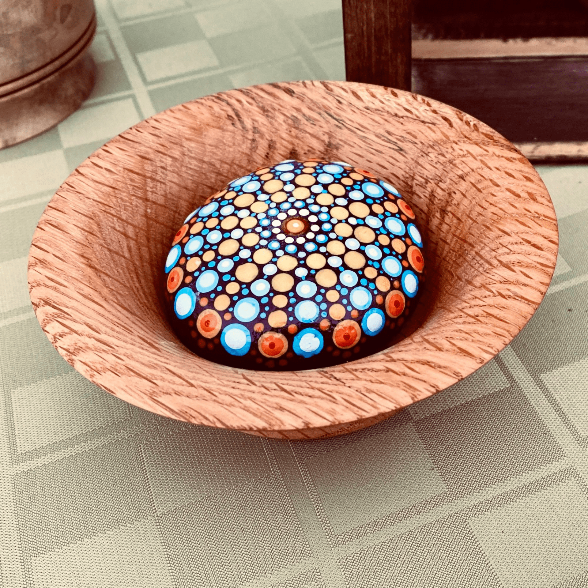 A colorful painted rock with intricate dot patterns inside a wooden bowl on a checkered tablecloth