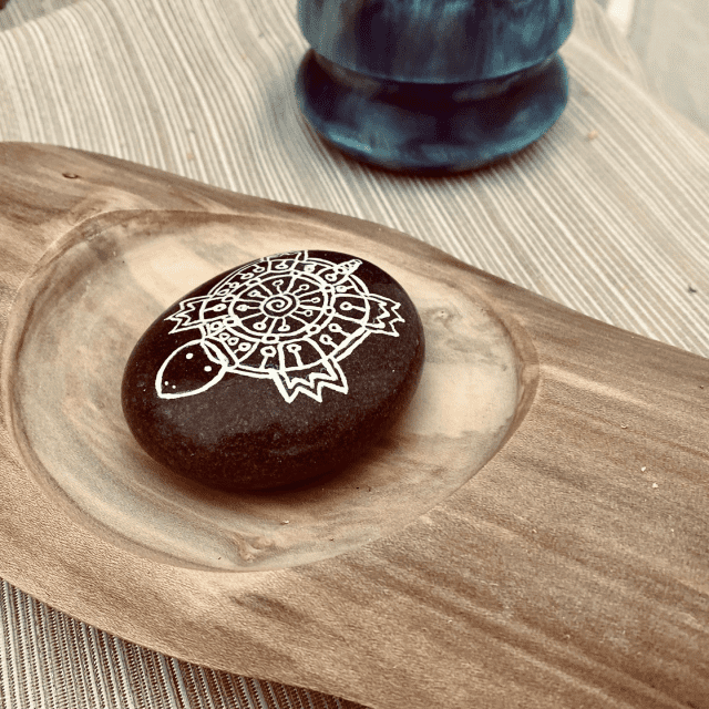 A painted stone with a mandala design resting on wooden platter, with a blue vase in the background A painted stone with a mandala design resting on wooden platter, with a blue vase in the background